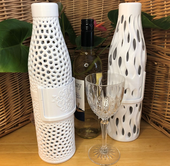 Two intricately designed, white 3D-printed wine bottle holders with unique cutout patterns, placed beside a wine bottle and a glass. A functional and stylish 3D print for wife who enjoys wine, making it a sophisticated accessory.
