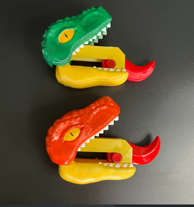 Two dinosaur-shaped knives in red and green, featuring T-Rex heads with teeth, a quirky and functional 3D print for kitchen enthusiasts.