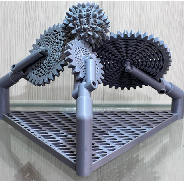 A 3D-printed gear system featuring multiple interlocking gears of various sizes, arranged on a triangular base with a grid-like platform. The gears are intricately designed, showcasing fine details and smooth rotational functionality. The structure is supported by angled rods, highlighting its mechanical complexity and aesthetic appeal.






