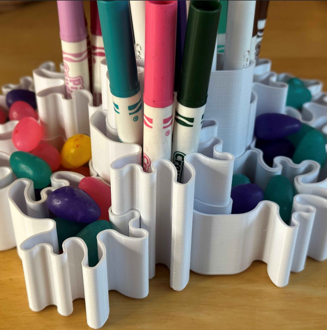 A white, wavy-designed desk organizer holding markers and jelly beans, placed on a wooden surface.
