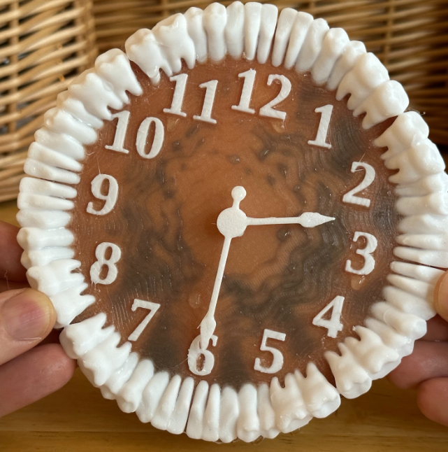 A unique 3D-printed clock with a ring of white teeth around the edge, placed against a wicker basket backdrop.
