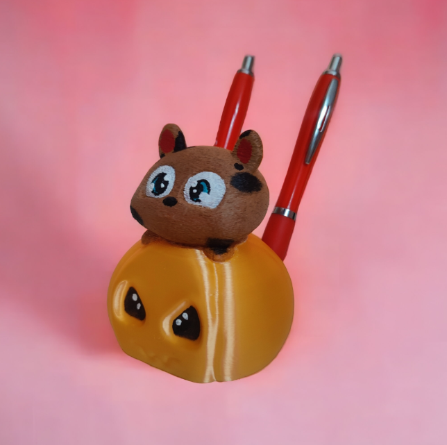 A yellow pumpkin-shaped 3D pen holder with a cute hamster on top, holding red pens on a pink background.
