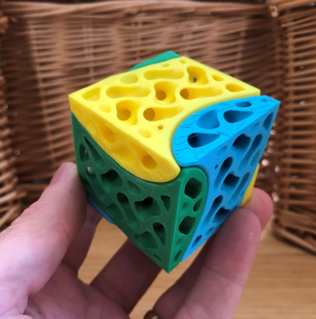  A vibrant multicolored 3D-printed interlocking cube, designed with intricate cutouts, being held in a hand.
