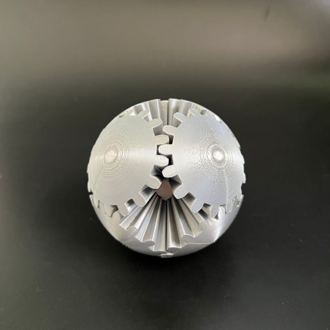 A silver spherical 3D-printed fidget toy with interlocking gears, resting on a black surface.
