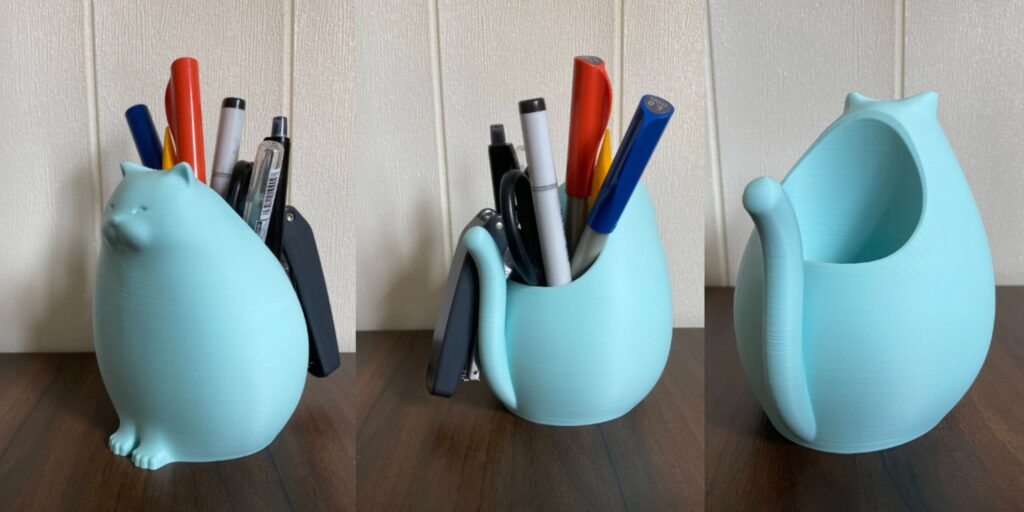  A pastel blue 3D-printed cat-shaped pencil holder, holding colorful pens and pencils on a wooden desk.
